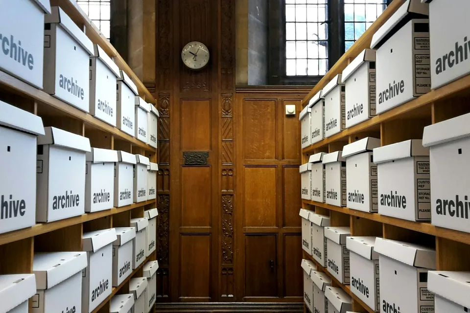 Corredor de arquivo com prateleiras de madeira repletas de caixas brancas identificadas como “archive”.