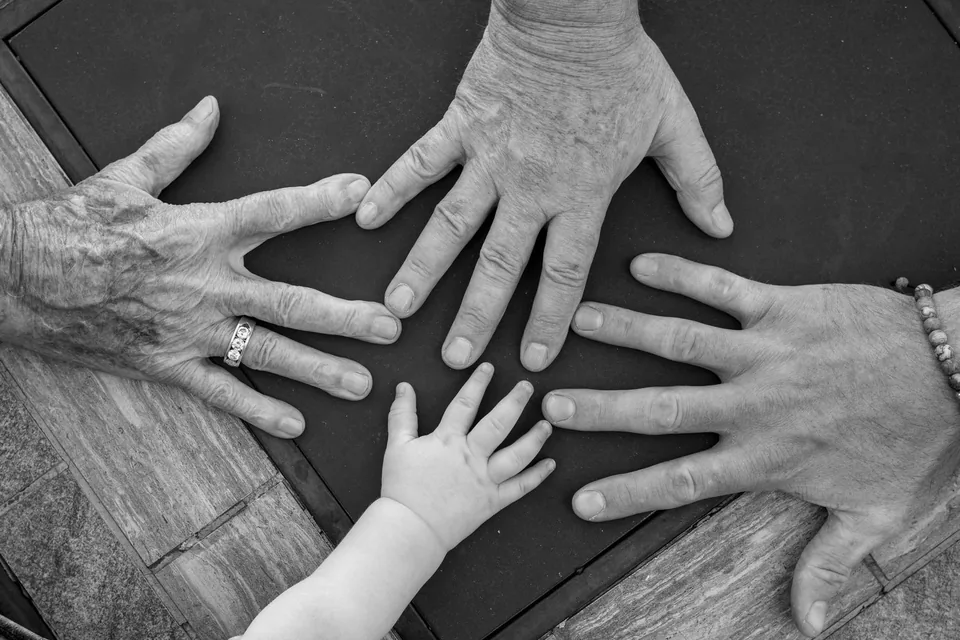 Mãos de quatro gerações, de uma criança a uma pessoa idosa, reunidas sobre uma mesa de madeira, em preto e branco.