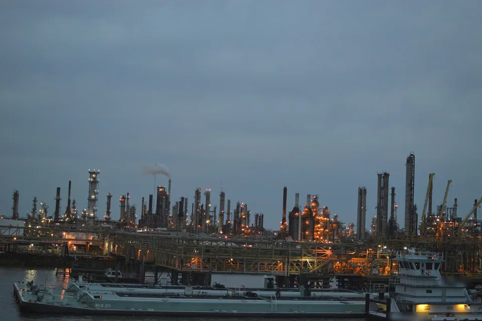 Complexo de refinaria iluminado ao entardecer, com barcaças no rio em primeiro plano.