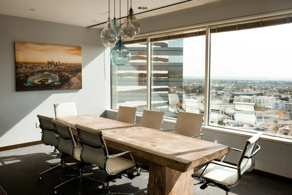 Sala de reuniao vazia com mesa de madeira clara, cadeiras brancas e vista da cidade pela janela.