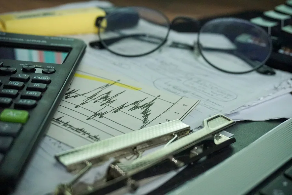 Mesa de trabalho com calculadora, óculos, papéis com gráficos e um grampeador, em tons escuros.