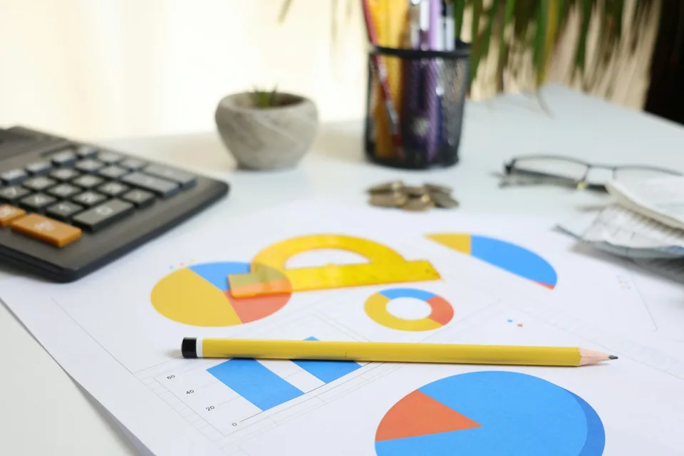Mesa de escritório com calculadora, gráficos impressos em papel e um lápis amarelo.