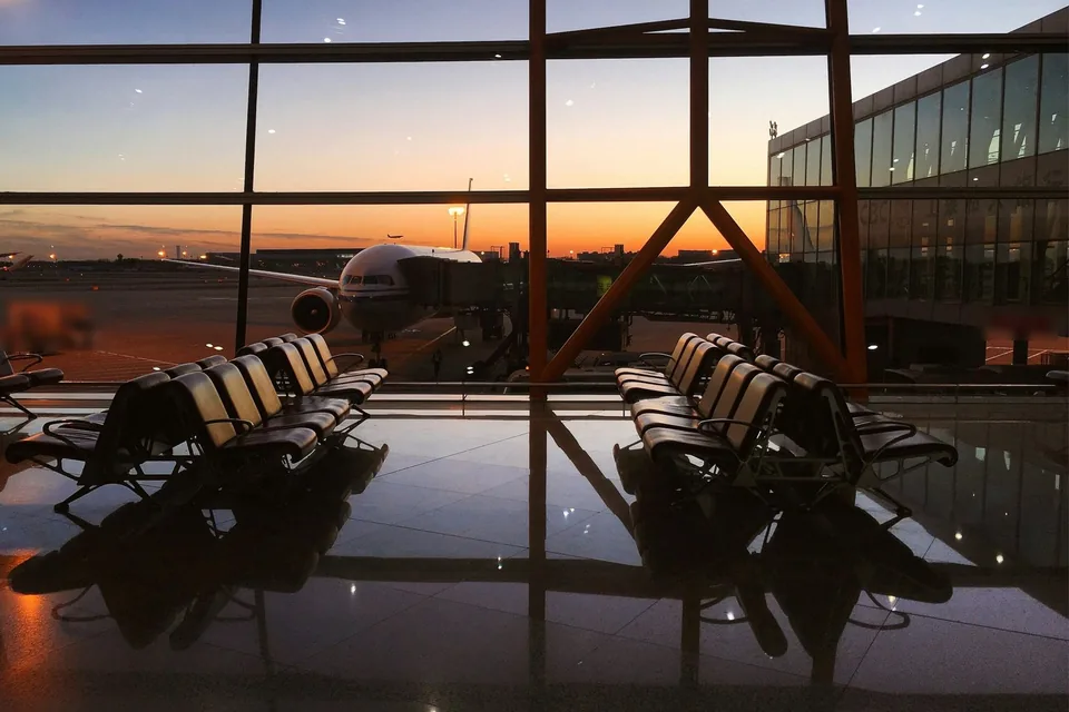 Sala de embarque vazia de um aeroporto ao pôr do sol, com fileiras de cadeiras diante da janela e um avião ao fundo.